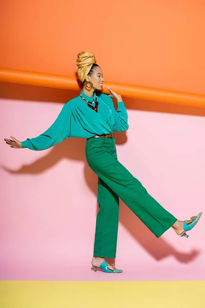 Comprimento total de mulher americana africana alegre em roupa elegante sorrindo enquanto segurando laranja rolou papel de parede no fundo rosa — Fotografia de Stock