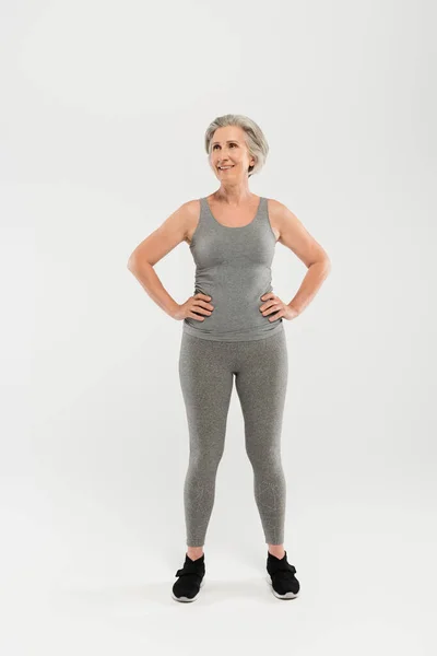 Volle Länge der glücklichen und älteren Frau in Sportbekleidung stehend mit den Händen auf den Hüften auf grau — Stockfoto