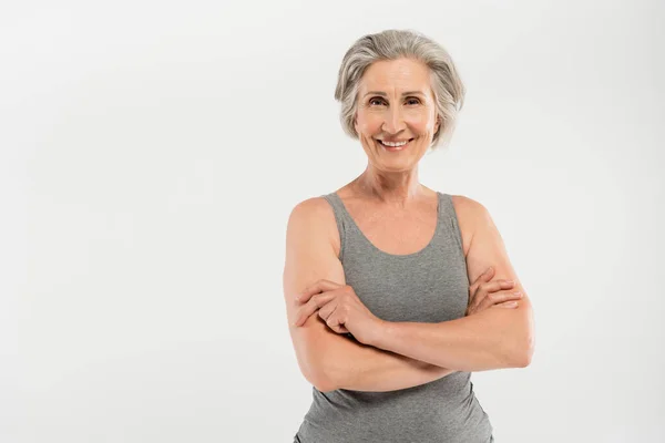 Mujer feliz y mayor en ropa deportiva de pie con los brazos cruzados aislados en gris - foto de stock
