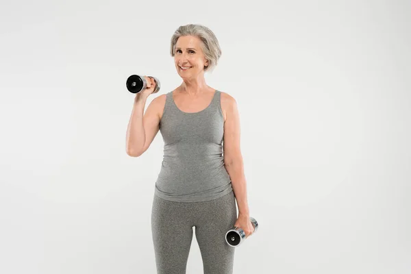 Mujer mayor positiva con pelo gris haciendo ejercicio con mancuernas aisladas en gris - foto de stock