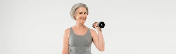 Cheerful senior woman with grey hair exercising with dumbbells isolated on grey, banner — Stock Photo