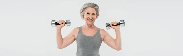 Mulher idosa alegre com cabelos grisalhos exercitando com halteres isolados em cinza, banner — Fotografia de Stock
