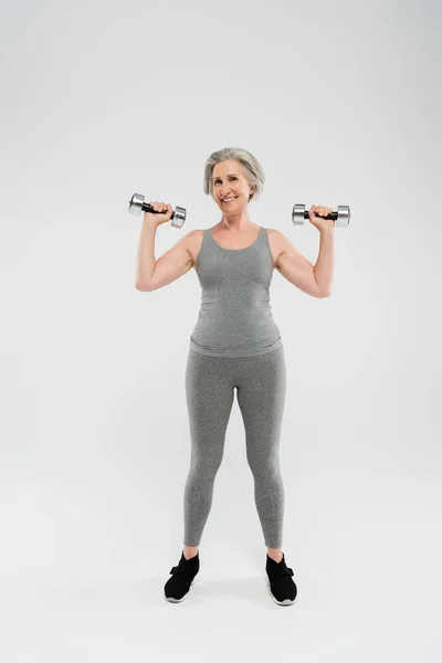 Full length of happy senior woman in tight sportswear exercising with dumbbells on grey — Stock Photo