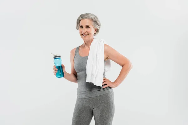 Щаслива старша жінка з білим рушником на плечі тримає спортивну пляшку з водою ізольовано на сірому — стокове фото