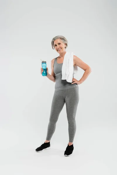 Pleine longueur de femme âgée gaie avec serviette blanche sur l'épaule tenant bouteille de sport avec de l'eau sur gris — Photo de stock