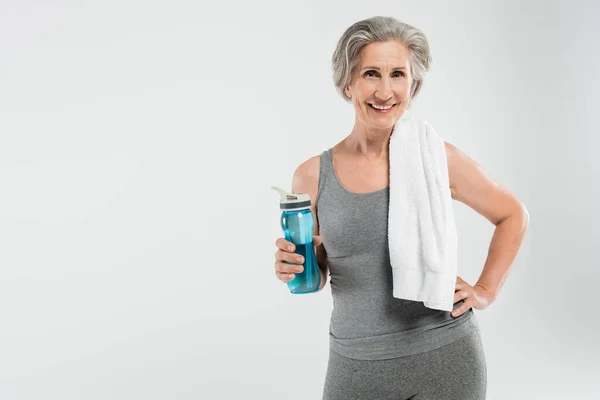Mujer mayor llena de alegría con toalla blanca en el hombro sosteniendo botella deportiva con agua aislada en gris - foto de stock