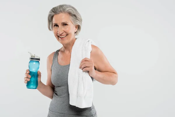 Mujer mayor complacida con toalla blanca en el hombro sosteniendo botella deportiva con agua aislada en gris — Stock Photo