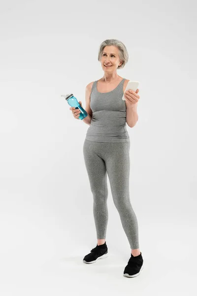 Piena lunghezza di felice donna anziana in abbigliamento sportivo con bottiglia sportiva con acqua e smartphone su grigio — Foto stock