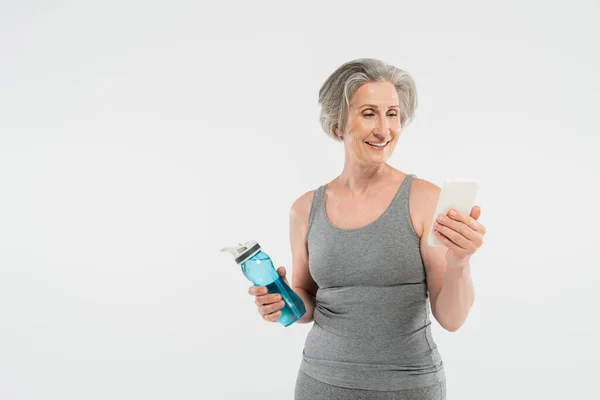 Щаслива старша жінка в спортивному одязі тримає спортивну пляшку з водою і використовує смартфон ізольовано на сірому — стокове фото