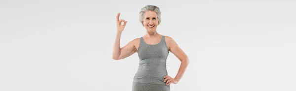 Mujer alegre y jubilada en ropa deportiva posando con la mano en la cadera y mostrando bien aislado en gris, bandera - foto de stock