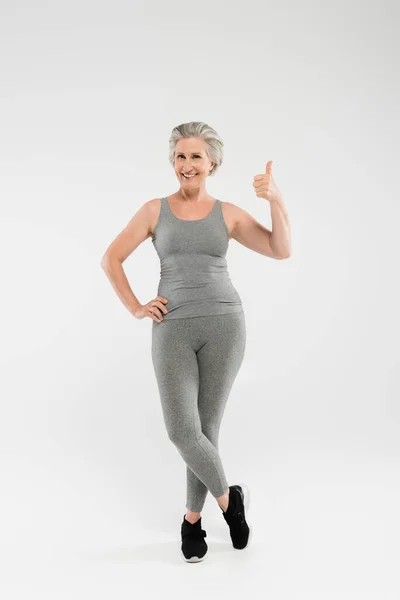 Повна довжина весела і пенсіонерка в спортивному одязі позує з рукою на стегні і показує, як на сірому — стокове фото