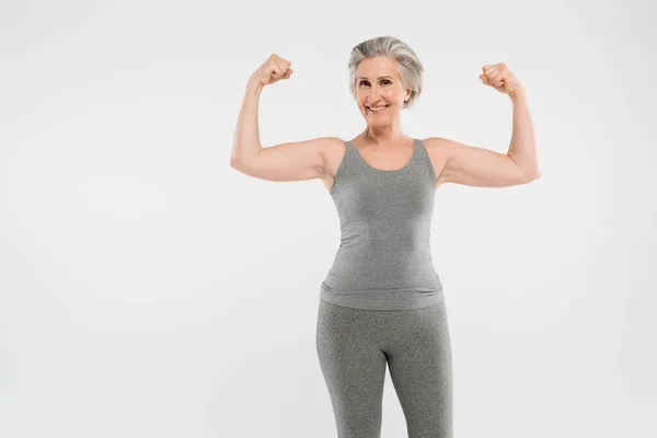 Cheerful and retired woman in sportswear showing muscles isolated on grey — Stock Photo
