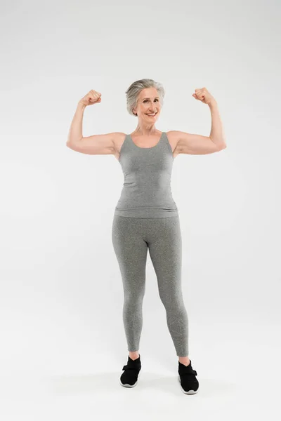 Longitud completa de la mujer alegre y jubilada en ropa deportiva que muestra los músculos en gris - foto de stock
