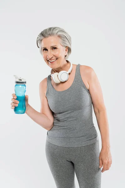Glückliche Seniorin mit drahtlosen Kopfhörern, die Sportflasche isoliert auf grau gehalten — Stockfoto