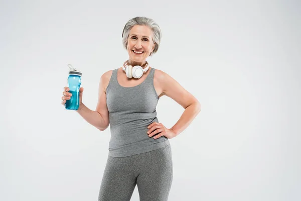 Mujer mayor feliz con auriculares inalámbricos sosteniendo botella deportiva y de pie con la mano en la cadera aislado en gris - foto de stock