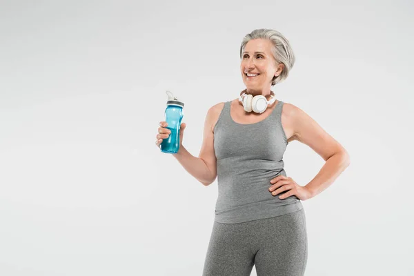 Mujer mayor despreocupada con auriculares inalámbricos que sostienen la botella deportiva y de pie con la mano en la cadera aislada en gris — Stock Photo