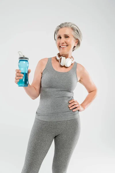 Улыбающаяся пожилая женщина с беспроводными наушниками держа спортивную бутылку и стоя с рукой на бедре изолированы на сером — стоковое фото