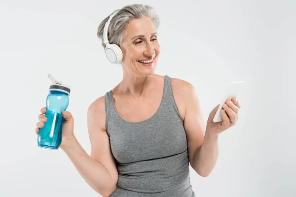 Joyful senior woman listening music in wireless headphones while holding sports bottle and smartphone isolated on grey — Stock Photo