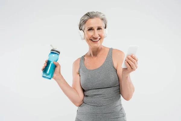 Carefree senior woman listening music in wireless headphones while holding sports bottle and smartphone isolated on grey — Stock Photo