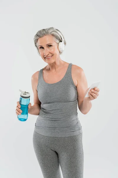 Fröhliche Seniorin hört Musik in drahtlosen Kopfhörern, während sie Sportflasche und Smartphone isoliert auf grau hält — Stockfoto