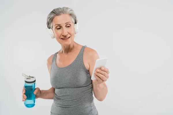 Щаслива старша жінка слухає музику в бездротових навушниках, тримаючи спортивну пляшку і смартфон ізольовано на сірому — стокове фото