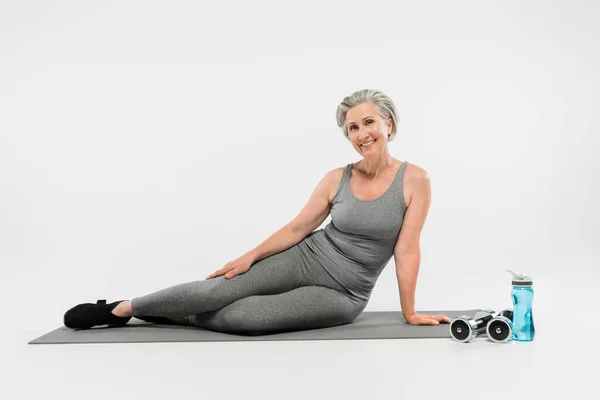 Pleine longueur de femme âgée heureuse en vêtements de sport assis sur tapis de fitness près de bouteille de sport et haltères sur gris — Photo de stock