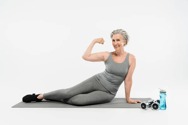 Comprimento total de mulher idosa alegre sentado no tapete de fitness perto de garrafa de esportes e halteres ao mostrar o músculo no cinza — Fotografia de Stock