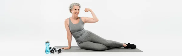 Pleine longueur de femme âgée gaie assis sur tapis de fitness près de bouteille de sport et haltères tout en montrant le muscle sur gris, bannière — Photo de stock