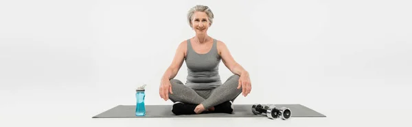 Longitud completa de la mujer mayor feliz sentada en postura del yoga en la estera cerca de las mancuernas y de la botella del deporte en gris, bandera - foto de stock