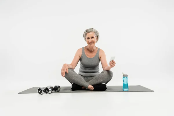 Pleine longueur de femme âgée gaie assis dans la pose de yoga sur le tapis près des haltères et en utilisant smartphone sur gris — Photo de stock