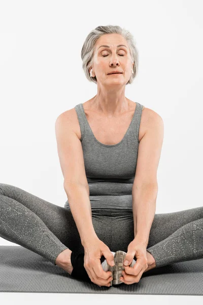 Senior woman with wireless earphones sitting in yoga pose on mat isolated on grey — Stock Photo