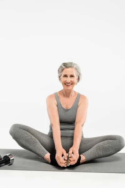 Полная длина счастливой пожилой женщины с беспроводными наушниками, сидящей в позе йоги на сером — стоковое фото