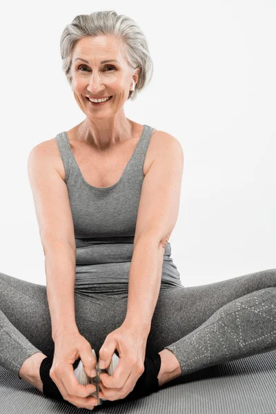 Mulher sênior positivo com fones de ouvido sem fio sentado em pose de ioga no tapete cinza isolado — Fotografia de Stock