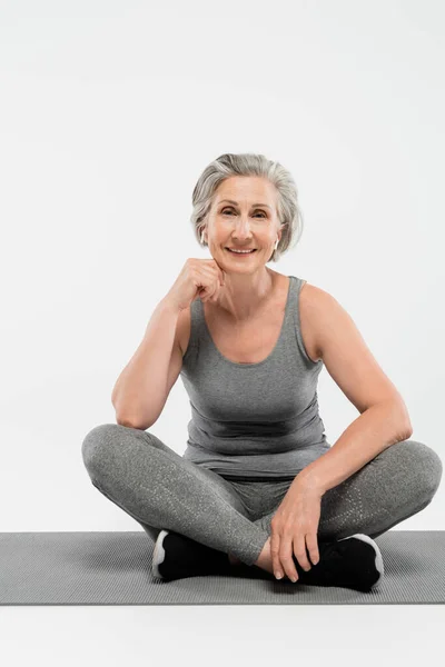 Повна довжина задоволеної старшої жінки з бездротовими навушниками, що сидять в позі йоги на сірому — стокове фото