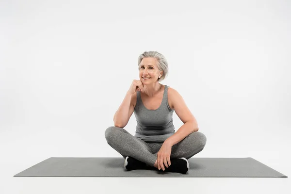 Volle Länge der zufriedenen Seniorin in drahtlosen Kopfhörern, die Musik hört und in Yoga-Pose auf grau sitzt — Stockfoto