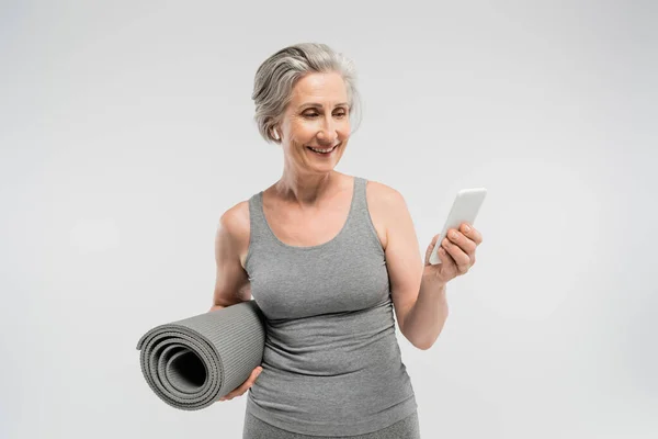 Весела старша жінка в бездротових навушниках, що тримає фітнес-матер і смартфон ізольовані на сірому — стокове фото