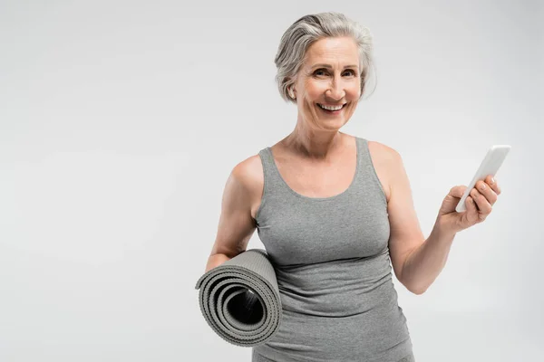 Donna anziana spensierata con auricolari wireless con stuoia fitness e smartphone isolati su grigio — Foto stock
