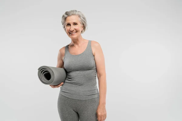 Unbekümmerte Seniorin in Sportbekleidung hält Fitnessmatte in der Hand und lächelt vereinzelt auf grau — Stockfoto