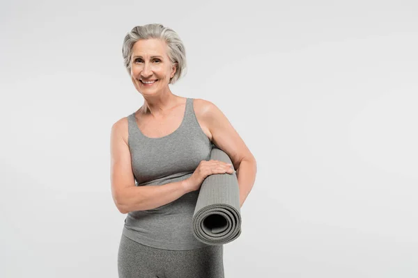 Весёлая пожилая женщина в спортивной одежде держит фитнес-мат и улыбается изолированно на сером — стоковое фото