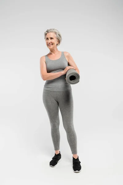 Piena lunghezza della donna anziana allegra in abbigliamento sportivo in possesso di tappetino fitness e in piedi sul grigio — Foto stock