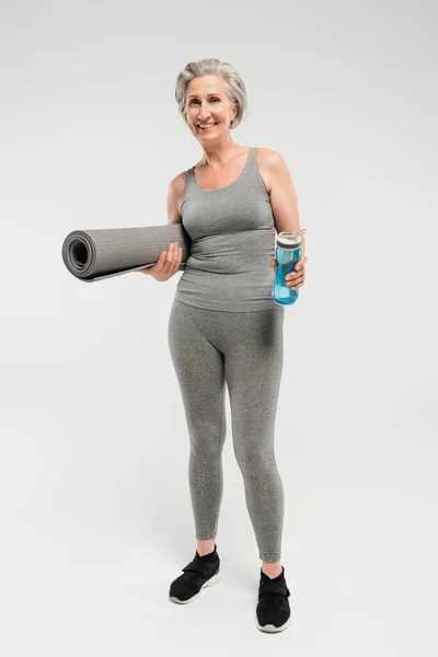 Full length of cheerful senior woman holding fitness mat and sports bottle with water on grey — Stock Photo