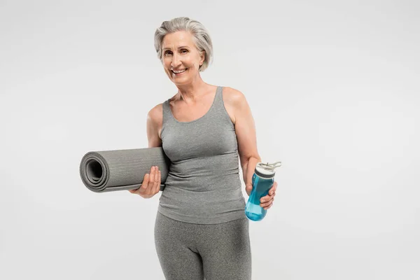 Веселая пожилая женщина держит фитнес мат и спортивную бутылку с водой, изолированной на сером — стоковое фото