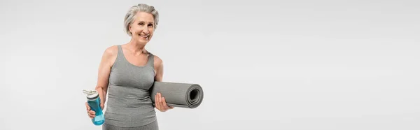 Alegre anciana sosteniendo alfombra de fitness y botella de deporte con agua aislada en gris, pancarta - foto de stock