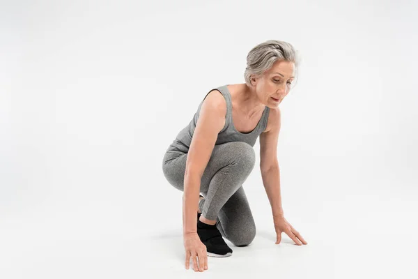 Full length of senior woman in sportswear standing in start position before run on grey background — Stock Photo