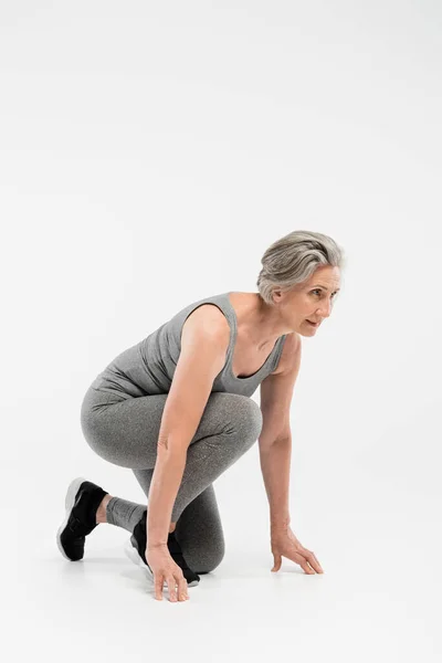 Volle Länge der Rentnerin in Sportbekleidung, die vor dem Lauf auf grauem Hintergrund in der Startposition steht — Stockfoto