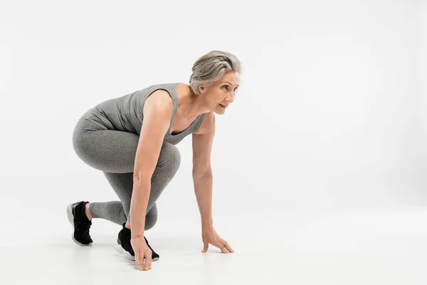 Full length of senior woman in leggings and tank top standing in start position before run on grey background — Stock Photo