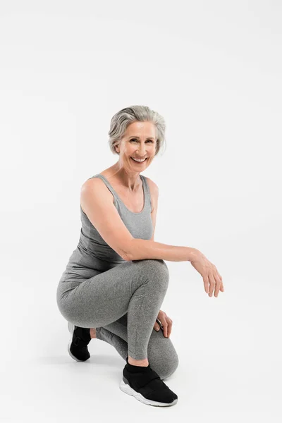 Pleine longueur de femme âgée gaie en vêtements de sport souriant tout en étant assis sur gris — Photo de stock