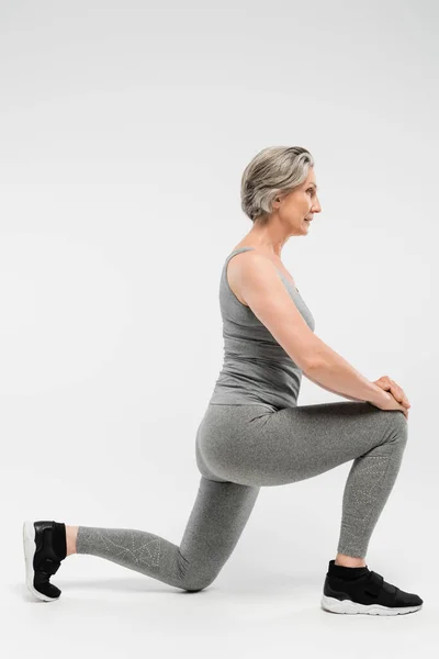 Full length of senior woman in sportswear doing lunges on grey — Stock Photo