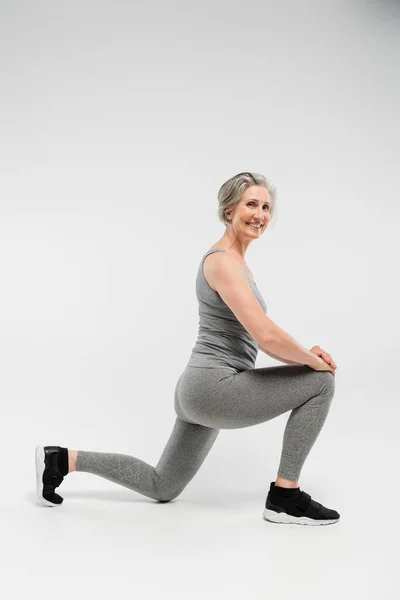 Pleine longueur de femme âgée heureuse en vêtements de sport faisant des fentes et souriant sur gris — Photo de stock