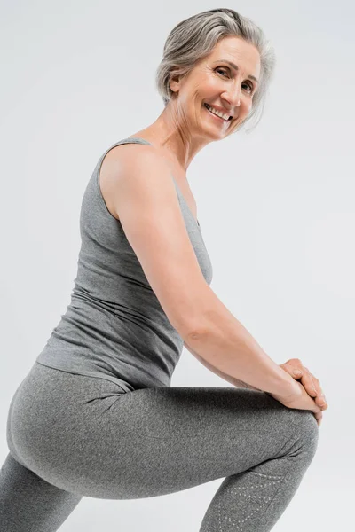 Mulher sênior feliz em sportswear fazendo lunges e sorrindo isolado em cinza — Fotografia de Stock